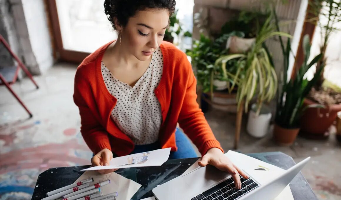 Young beautiful woman with dark curly hair sitting at the table thoughtfully working on laptop with fashion illustration in hand while spending time in modern cozy workshop with big windows
