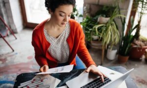 Young beautiful woman with dark curly hair sitting at the table thoughtfully working on laptop with fashion illustration in hand while spending time in modern cozy workshop with big windows