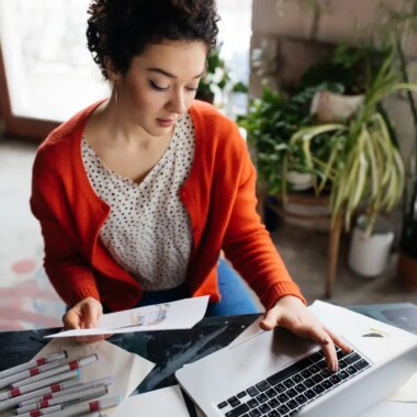 Young beautiful woman with dark curly hair sitting at the table thoughtfully working on laptop with fashion illustration in hand while spending time in modern cozy workshop with big windows