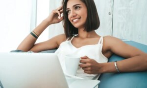 Portrait of beautiful smiling woman dressed in white pajamas. Carefree model sitting on soft bag chair and using laptop.