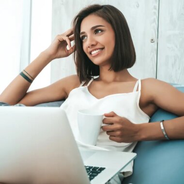 Portrait of beautiful smiling woman dressed in white pajamas. Carefree model sitting on soft bag chair and using laptop.