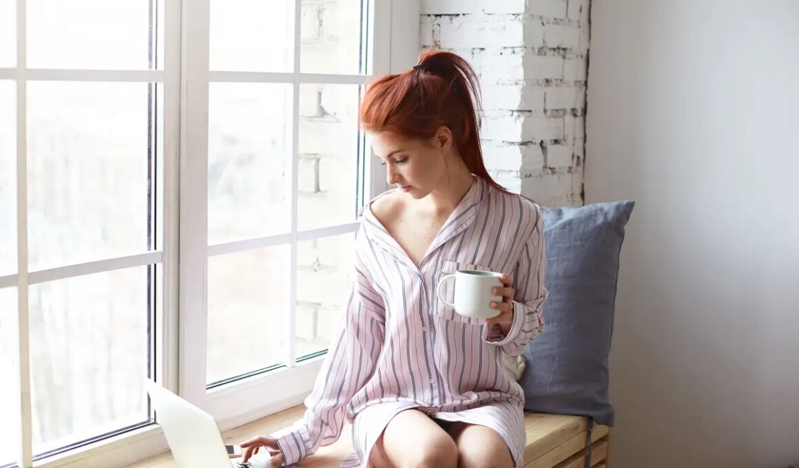 Modern technology, leisure and communication concept. Pretty redhead girl with ponytail sitting on windowsill with cup of hot drink, using high speed internet connection on generic laptop computer