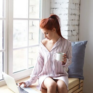 Modern technology, leisure and communication concept. Pretty redhead girl with ponytail sitting on windowsill with cup of hot drink, using high speed internet connection on generic laptop computer
