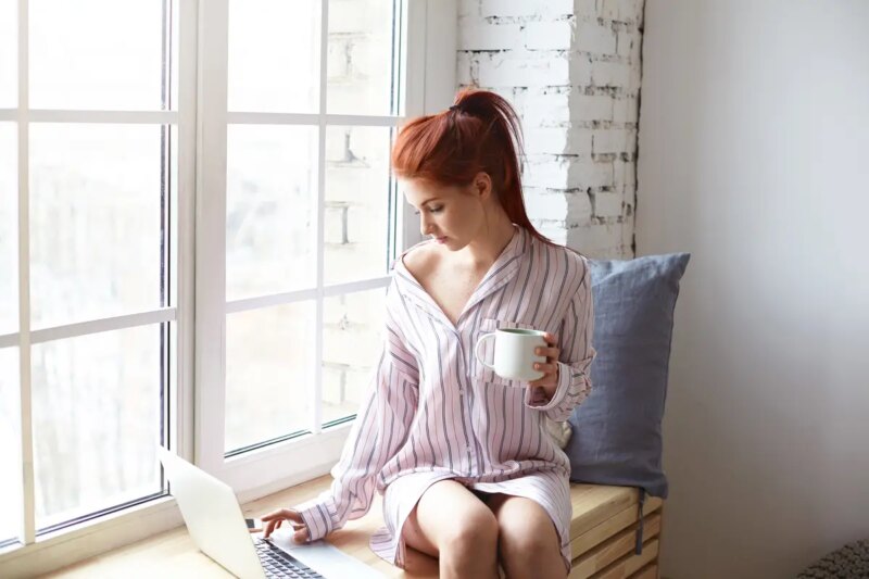 Modern technology, leisure and communication concept. Pretty redhead girl with ponytail sitting on windowsill with cup of hot drink, using high speed internet connection on generic laptop computer