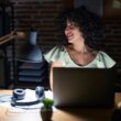 Young brunette woman with curly hair working at the office at night looking away to side with smile on face natural expression laughing confident