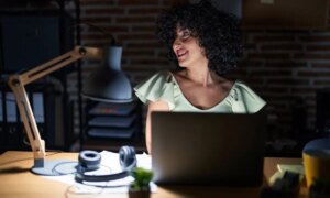 Young brunette woman with curly hair working at the office at night looking away to side with smile on face natural expression laughing confident