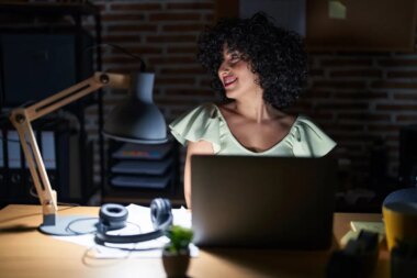 Young brunette woman with curly hair working at the office at night looking away to side with smile on face natural expression laughing confident